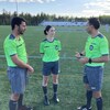 Trois personnes en discussion sur une surface synthétique avec des joueurs de soccer en arrière-plan