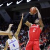 Le joueur de basketball canadien, en rouge, s'élance dans les airs pour tenter un tir. Il tient le ballon à deux mains devant deux adversaires de la République dominicaine, en blanc. 