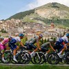 Un peloton de cyclistes roule en contrebas d'un village pittoresque à flanc de montagne. 