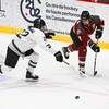 Deux joueuses de hockey d'équipes adverses tentent de maîtriser une rondelle, elles se trouvent à gauche du filet.
