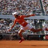 Le joueur de tennis serbe, avec un maillot orange et un short et une casquette blanche, saute pour frapper la balle du revers sur la terre battue. 
