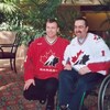 Martin Brodeur est accroupi à côté de Pierre Pichette, assis dans un fauteuil roulant. Les deux hommes portent un chandail de l'équipe canadienne de hockey.