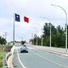 Des voitures circulent sur une route surplombée par un drapeau de l'Acadie sur un haut mât.