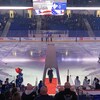 Des joueurs de hockey évoluent sur une patinoire et des spectateurs sont debout dans les gradins pendant qu'une femme est sur un tapis rouge sur la glace.