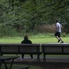 Une personne court dans un parc et une autre la regarde, assise sur un banc.