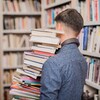 Un jeune homme vu de dos, portant de nombreux livres en équilibre, dans une bibliothèque.