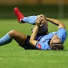 Une joueuse tient son genou droit entre ses mains, couchée sur son dos en douleur sur la pelouse d'un terrain de soccer. 