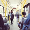 Une adolescente est debout dans l'allée pendant les trajets en bus. D'autres adultes sont assis de dos, le regard vers l'avant du bus.