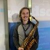 Un jeune homme dans la vingtaine, souriant, tient un saxophone et est debout devant une affiche sur laquelle est écrit Windsor.
