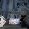 Deux personnes tiennent un banderole demandant de refuter la doctrine debout sur les marches d'une cathédrale.