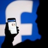 Une personne à contre-jour tient un téléphone ouvert sur Facebook. En fond, le f de Facebook sur un grand écran.