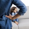 On voit une petite fille recroquevillée sur un sofa avec la silhouette d'un parent en avant-plan.