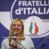 Portrait cadré d'une femme sur une scène, derrière un lutrin, qui tient un micro dans ses mains et sourit en regardant au loin. Derrière elle, le logo de son parti politique, une flamme tricolore, hérité du mouvement social italien de 1946. 