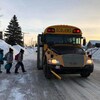 Dans un quartier résidentiel l'hiver, trois enfants montent à bord d’un autobus scolaire.