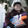 Un garçon de 10 ans, vêtu d'un manteau d'hiver et tenant un pistolet à eau sous le bras, sourit depuis l'extérieur d'une maison, décorée pour le temps des Fêtes. 