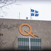 Gros plan sur l'enseigne lumineuse du logo d'Hydro-Québec installée au sommet du bâtiment du siège social de la société d'État près d'un drapeau du Québec qui flotte au vent.