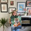 Une jeune femme aux cheveux longs, entourée de ses oeuvres d'arts, tient dans ses mains un tigre qu'elle a peint.