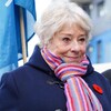Une femme aux cheveux blanc à l'extérieur. Elle porte un foulard multicolore, un manteau en lainage bleu et un coquelicot.