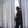 Une femme regarde l'extérieur par l'ouverture des rideaux, debout devant une fenêtre. 