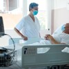 Une infirmière masquée parle à un patient alité dans une chambre d'hôpital.