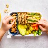 Vue aérienne d'une boîte à lunch compartimentée remplie d'un sandwich, de bâtonnets de carotte et de concombre, d'un demi-avocat, de raisins, de biscuits santé et de noix, avec une banane posée à côté. 