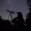 Une personne assise à côté de son télescope sous un ciel d'étoiles. 