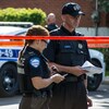 Une policière et un policier se tiennent debout derrière un bandeau de sécurité sur une scène de crime, devant une autopatrouille. 