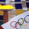 Un drapeau blanc paré des anneaux olympiques flotte devant la vasque de bronze où brûle la flamme olympique. 