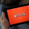 L'écran d'une tablette, tenue par un homme dont on aperçoit les pieds en arrière plan, affiche le logo de Netflix.