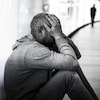 Une homme visiblement triste est assis par terre. Il tient son visage dans ses mains. Il est habillé d'un chandail à capuche et d'un pantalon en jean.