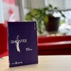 Un livre violet sur une petite table, avec des plantes et des fauteuils rouge en arrière-plan. 