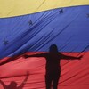 La silhouette d'une femme, debout, les bras étendus, se profile derrière le drapeau du Venezuela.