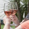 Les mains de deux personnes qui se font un toast avec un verre de vin rosé. Elles sont dehors l'été et il y a une bouteille sur la table.