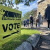 Une affiche indiquant la direction d'un bureau de vote au premier plan et une file d'attente en arrière-plan.