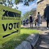 Une affiche indiquant la direction d'un bureau de vote en avant-plan et une file d'attente en arrière-plan.
