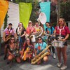 On aperçoit ici les sept membres du Burning BRASs Band, toutes des femmes. La photo a été prise à l'extérieur, en été. L'ambiance est festive. Plusieurs portent des couleurs tropicales ou même des colliers hawaïens. 