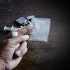 Une main qui tient de l'argent et de la poudre d'héroïne dans un sac. Une seringue, un médicament, une cigarette et un briquet sont déposés sur une table en arrière-plan.