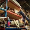 Une femme sur une échelle tient un meuble en bois sur une étagère dans un grand entrepôt.
