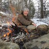 Francine Ouellette, souriante, assise au bord d'un feu dans une forêt.