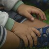 Des mains et un pied d'un jeune enfant à la peau foncée. L'enfant porte des vêtements de coton ouaté et des chaussures de sport neuves. Son chandail comporte une minuscule tache rougeâtre sur la manche. Il est assit dans l'herbe.