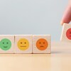 Gros plan de petits cubes en bois sur lesquels se trouvent des symboles qui représentent différentes émotions. Une main prend un cube sur lequel se trouve un symbole d'une émotion négative.