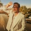 Un homme avec une barbe grise et des lunettes de soleil teintées orange pose avec une main levée pour se protéger les yeux du soleil. Il porte une veste de tailleur crème décontractée et une chemise en maille. L'arrière-plan montre une route en montée bordée d'arbres à Los Angeles, en Californie.