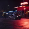 De nuit, le parking d'un motel où un homme tire sur une voiture, éclairée au néon.