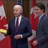 Joe Biden pose à côté de Justin Trudeau devant des drapeaux canadiens et américains. Il tient dans sa main une boîte où on peut lire le mot Peace.