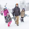 Deux adultes et quatre enfants marchent dans la neige en portant leurs skis et leurs planches à neige.