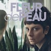 Devant une fenêtre et une plante verte, une femme détourne le regard vers la caméra.