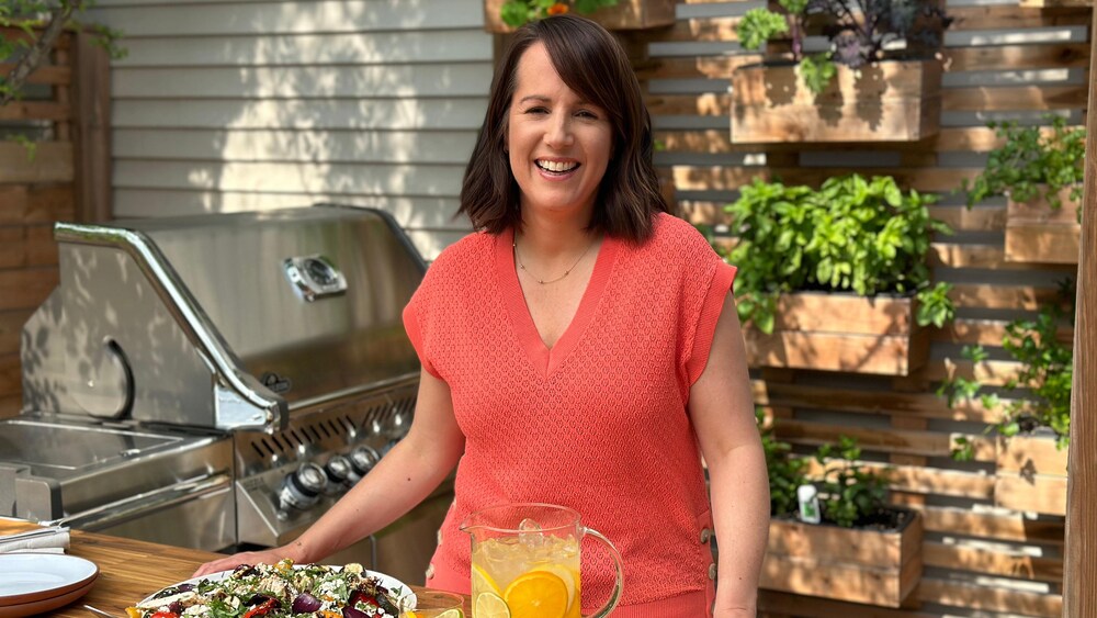 La nutritionniste Geneviève O’Gleman est sur la terrasse, près d’un barbecue et derrière une table sur laquelle se trouve une assiette de poulet et légumes grillés à la grecque et une agua fresca aux agrumes. 