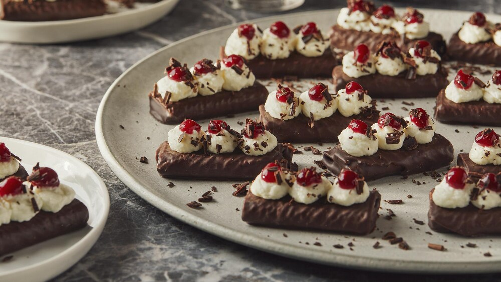 Une assiette remplie de crêpes dentelles au chocolat avec trois petites boules de crème fouettée dessus et trois petits points de confiture de framboises, le tout recouvert de copeaux de chocolat.