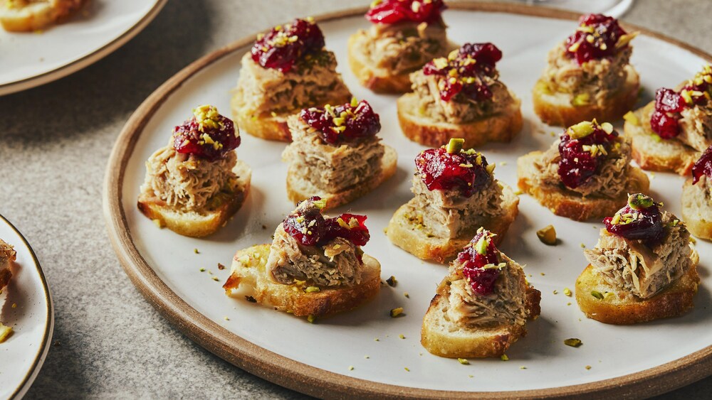 Dans une grande assiette, il y a une douzaine de canapés composés d’une tranche de baguette, un cube de rillettes, une compote de canneberges et des pistaches émiettées.