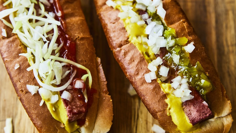 Il y a deux hot-dog sur une table. En arrière plan, il est possible de voir différents condiments.